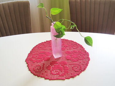 Rundes pinkes gehäkeltes Deckchen mit Rosenmuster auf weißem Tisch und rosa Vase