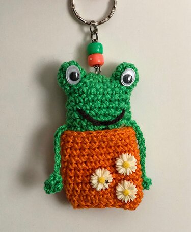 Gehäkelter Frosch-Schlüsselanhänger in Grün und Orange mit Wackelaugen, gesticktem Lächeln und drei kleinen Blüten auf dem Bauch