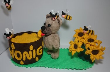 Gehäkelte Amigurumi-Szene: Teddybär am Honigtopf, umgeben von Bienen und Sonnenblumen auf grüner Matte.