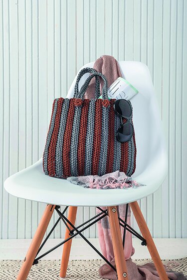 Gehäkelte Henkeltasche mit rostroten und graublauen Streifen auf weißem Stuhl mit Schal und Sonnenbrille