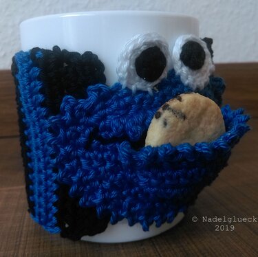 Weißer Becher mit handgehäkeltem blau-schwarzem Monster-Tassenwärmer mit kleinem Keksfach