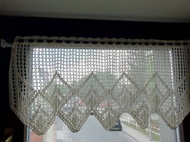 Hellfarbene, gehäkelte Scheibengardine mit filigranem Muster und geschwungener Unterkante, an einer Stange vor einem Fenster.