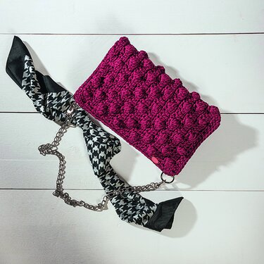 Bordeauxrote gehäkelte Clutch mit Noppenmuster, silberner Gliederkette und schwarz-weißem Tuch auf hellem Holzhintergrund.