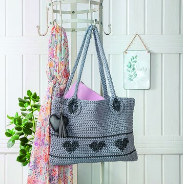 Gehäkelte graue Henkeltasche mit dunkelgrauen Herzen und herzförmigem Anhänger, an Garderobenhaken hängend