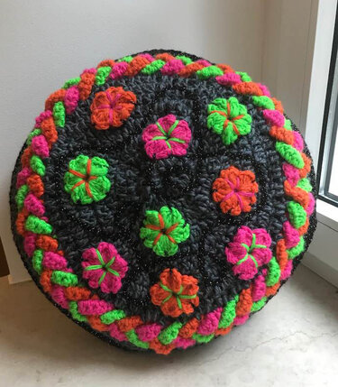 Handgehäkeltes, rundes Kissen aus dunklem Garn mit feinem Glitzereffekt, bestückt mit bunten, plastischen Blumenapplikationen in Neonrosa, Neonorange und Neongrün, umgeben von einem geflochtenen Rand.