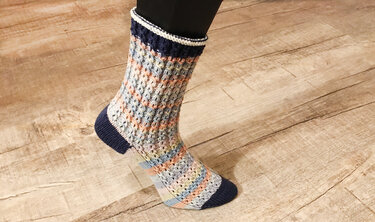 Mehrfarbiger gestrickter Strumpf mit Wabenmuster in Pastelltönen und dunkelblauen Akzenten auf einem Fuß auf Holzboden.