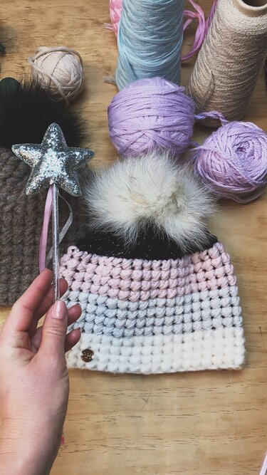 Gehäkelte Babymütze in Weiß, Rosa und Grau mit großem Kunstfellbommel; Garnknäuel im Hintergrund und Hand am Rand