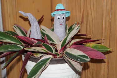 Weiß-graue gehäkelte Amigurumi-Figur mit türkisfarbenem Hut in weißem Blumentopf mit rosa-grün-weiß gestreiften Blättern vor Holzpaneel
