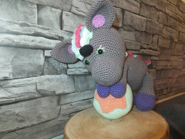 Grau gehäkeltes Einhorn mit pastellfarbener Mähne, violetten Hufen und grünen Augen auf Holzscheibe vor Steinwand