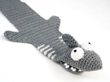 Gray crocheted shark bookmark with raised head, white eyes and teeth on white background