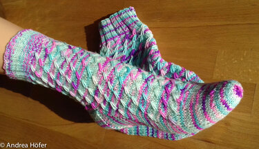 Gestrickte Socken mit Wabenmuster in Mint-, Pink- und Lilatönen; eine Socke am Fuß, eine liegt daneben.