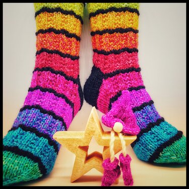 Nahaufnahme gestrickter mehrfarbiger Socken mit breiten Ringeln und schwarzen Trennlinien, davor Holzstern und kleines gestricktes Mützchen
