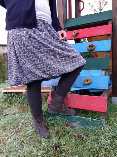 Frau mit grauem, gestreiftem Rock, dunklen Strumpfhosen und braunen Stiefeletten vor buntem Spielgerät