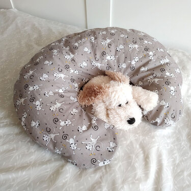 Graues U‑förmiges Kissen mit Affenmuster und Plüschhund auf hellem Bett.