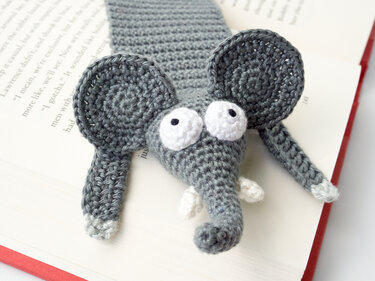 Grey crocheted elephant bookmark with white round eyes peeking from an open book.