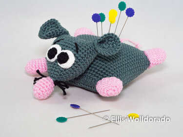 Handgehäkeltes Maus-Nadelkissen in Grau mit rosa Details, bestückt mit bunten Stecknadeln auf hellem Hintergrund.