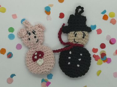 Gehäkelte Glücksbringer: rosafarbenes Schweinchen mit Pilz und schwarzer Schornsteinfeger, verbunden durch rote Schnur, auf weißem Hintergrund mit Konfetti.