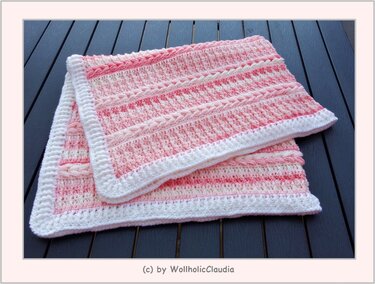 Rosa-weiße gehäkelte Babydecke mit Zopfmuster und weißer Umrandung, auf dunklem Tisch liegend