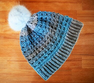 Gehäkelte blau-graue Damenmütze mit Rippbündchen und weißem Bommel auf Holzuntergrund