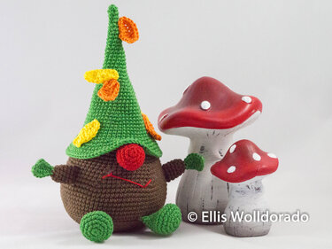 Braunes, rundes Amigurumi mit grünem Spitzhut und zwei rot-weiß gepunkteten Pilzen