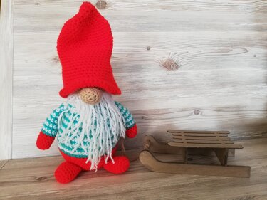 Gehäkelter Weihnachtswichtel mit roter Mütze, weißem Bart und Holzschlitten vor hellem Holz.