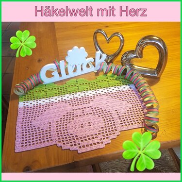 Gehäkeltes rosa Deckchen mit Herzmuster und grünem Rand auf Holztisch, daneben Dekoherzen und Schriftzug 'Glück'.