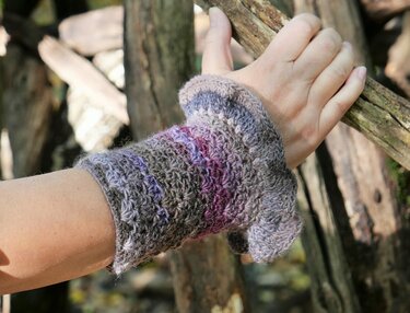 Nahaufnahme einer fingerlosen Armstulpe mit Muschelmuster und Daumenöffnung in Grau‑ und Lilatönen