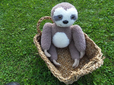 Amigurumi-Faultier mit braun-weißem Fell in einem Weidenkorb auf grüner Wiese