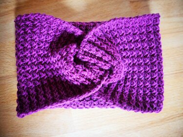 Nahaufnahme eines gehäkelten Stirnbands in Lila mit Twist-Knoten in der Mitte auf einer Holzoberfläche