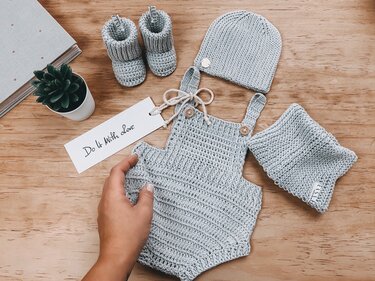 Hellgraues gehäkeltes Babyset mit Strampler, Mütze und Schühchen auf Holz, Hand hält Strampler