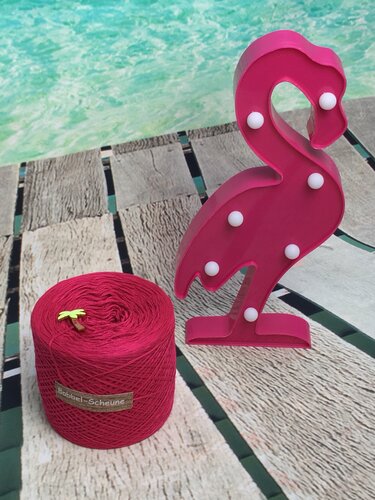 Roter Garnbobbel neben pinker Flamingo-Leuchte auf Holzplanken vor türkisfarbenem Wasser.