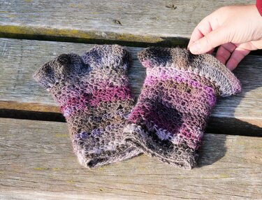 Zwei gehäkelte fingerlose Handschuhe in Grau-, Braun- und Violetttönen auf einer Holzoberfläche, eine Hand hält eine Stulpe.