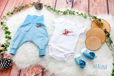 Baby-Body mit Hubschrauber-Applikation, gemusterte Pumphose und Babyschuhe auf flauschigem Untergrund.