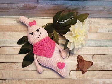 Hellrosa gehäkeltes Gespenst mit Schleife, pinkem Halstuch und Herz auf Holzhintergrund neben Blättern und weißer Blüte.