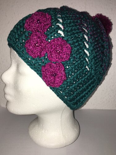 Dunkelgrüne gehäkelte Mütze mit vier pinken Blumenapplikationen, Perlen und Bommel auf Schaufensterkopf