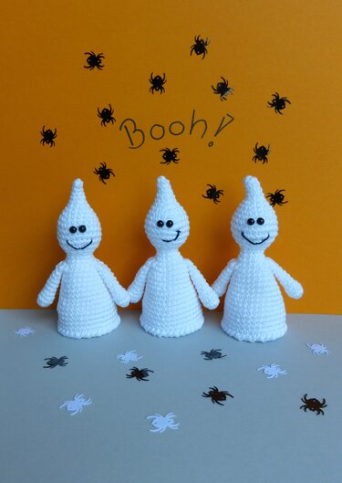 Drei weiße gehäkelte Geister mit lächelnden Gesichtern vor orangefarbenem Hintergrund mit kleinen Spinnen und dem Schriftzug „Booh!“, auf hellgrauem Untergrund mit Spinnenkonfetti.