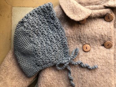 Gehäkelte graue Babymütze mit Bindebändern auf beigem Babymantel mit Holzknöpfen.