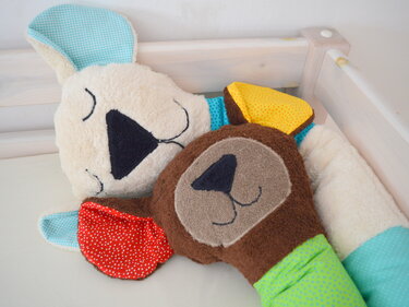 Zwei weiche, genähte Kuscheltiere (cremefarbener Hase und brauner Bär) mit bunten Stoffdetails im Babybett