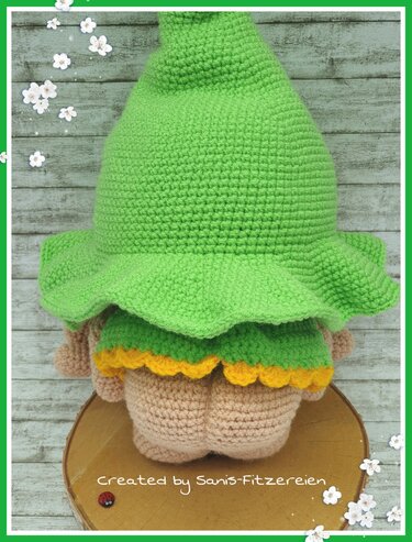 Rückansicht eines gehäkelten Amigurumi-Wichtels mit großem grünem Spitzhut, grüner Kleidung und gelbem Saum auf einer Holzscheibe.