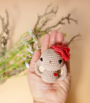 Kleiner beiger Amigurumi-Hase mit roter Blume am Kopf und kleinem roten Herz, in einer Hand gehalten