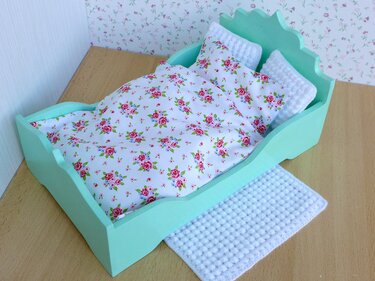 Mintgrünes Puppenbett mit weißer Bettwäsche im Rosenmuster, zwei gehäkelten Kissen und gehäkeltem Teppich auf Holzboden.