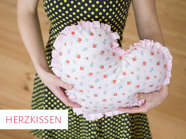 Eine Person hält ein rosa Herzkissen mit Blumenmuster und Rüschen.