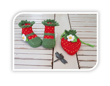 Red and green knitted baby booties with white flower and a strawberry-shaped knitted pouch on wooden slats