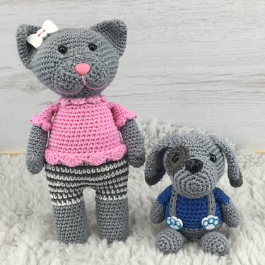 Gehäkelte graue Katze mit rosa Oberteil und gestreiften Beinen neben kleinem gehäkelten grauen Hund mit blauem Pulli auf hellem Teppich