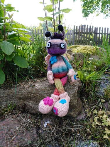 Buntes handgehäkeltes Amigurumi-Insekt mit großen Augen, Antennen und Blumenverzierung auf einem Stein im Garten