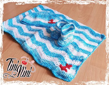 Gehäkeltes Baby-Set blau-weiß mit Wellenmuster, Mütze und Schühchen, rote Seestern- und Anker-Applikationen auf Holzboden.