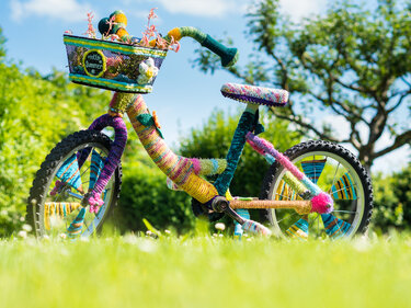 Bunt gestricktes Fahrrad mit bestricktem Korb auf grüner Wiese.