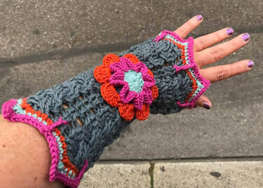 Nahaufnahme einer Hand mit einer grauen, gehäkelten, fingerlosen Armstulpe mit mehrfarbiger Blumenapplikation und bunten Rändern.
