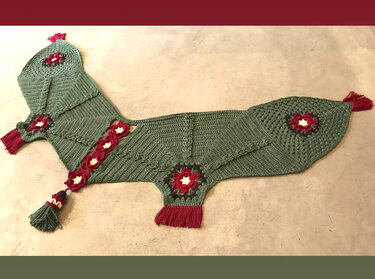 Grünes gehäkeltes Tuch mit roten Blütenapplikationen und Quasten auf hellem Untergrund.