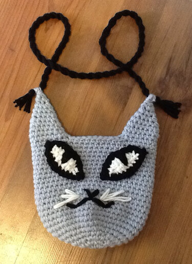 Hellgraue gehäkelte Umhängetasche in Katzenform mit schwarzem geflochtenem Trageband, applizierten Augen und weißen Schnurrhaaren.